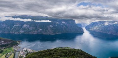 Norveç 'teki Aurlandsfjord' un nefes kesici görüntüsü, Stegastein bakış açısından çekildi. Aurlandsvangen 'in küçük kasabası fiyortun kıyısında, yüksek dağlar ve alçak asılı bulutlar dramatik bir zemin oluşturur. Turistler ziyarete gelir