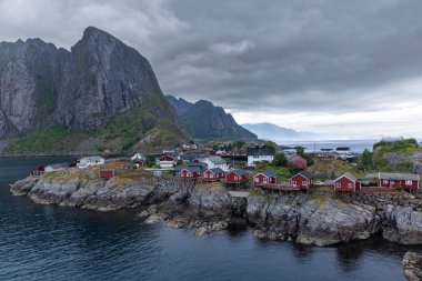 Norveç 'in Hamny kentindeki balıkçı köyünün manzarası. İkonik kırmızı rorbu kulübeleri kayalık çıkıntıların üzerine tünemişti. Bulutlu bir gökyüzünün altında dramatik dağlar vardı.. 