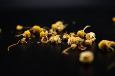 Koyu bir yüzeyde sarı merkezli kurumuş papatya çiçekleri ve soluk yapraklar, bitki çayını, rahatlamayı, doğal çareleri ve yatıştırıcı sağlık faydalarını sembolize ediyor..