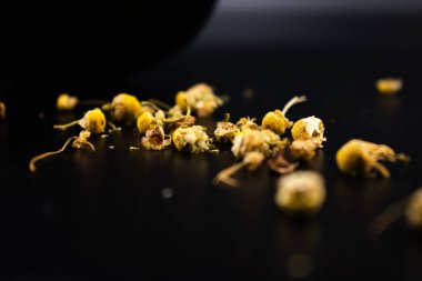Koyu bir yüzeyde sarı merkezli kurumuş papatya çiçekleri ve soluk yapraklar, bitki çayını, rahatlamayı, doğal çareleri ve yatıştırıcı sağlık faydalarını sembolize ediyor..