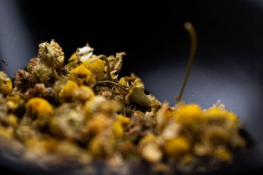 Koyu bir yüzeyde sarı merkezli kurumuş papatya çiçekleri ve soluk yapraklar, bitki çayını, rahatlamayı, doğal çareleri ve yatıştırıcı sağlık faydalarını sembolize ediyor..