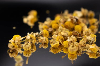 Koyu bir yüzeyde sarı merkezli kurumuş papatya çiçekleri ve soluk yapraklar, bitki çayını, rahatlamayı, doğal çareleri ve yatıştırıcı sağlık faydalarını sembolize ediyor..