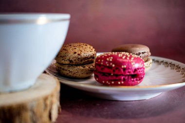 Koyu kahveyle doldurulmuş altın kaplamalı beyaz porselen bardak, bir tabak pembe ve kahverengi makaronun yanındaki ahşap bir bardak üzerinde serpiştirilir. Burgonya desenli arka plan hoş ve zarif bir dokunuş ekliyor..