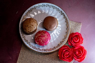 Çuval bezinin üzerindeki beyaz dekoratif tabak renkli badem kurabiyeleri içeriyor, samimi ve davetkar bir ortam yaratıyor. Yumuşak dokular ve ılık tonlar rahatlatıcı bir sabahı ve tatlı bir hoşgörünün zevkini çağrıştırıyor..