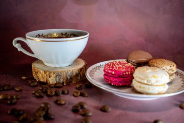 Beyaz kahve fincanı, ahşap altlık üzerinde altın işlemeli ve yanında renkli bir tabak makarna, kahve, pembe ve vanilya. Bordo bir yüzeye saçılmış kahve çekirdekleri sıcak ve davetkar bir manzara yaratır..