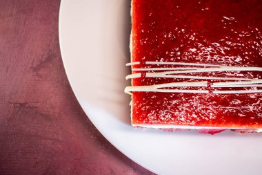 Bir dilim katmanlı sünger keki kremalı ve üzeri parlak kırmızı böğürtlenli jöleli, üzeri de beyaz çikolata telleriyle kaplı. Canlı renkler ve zengin dokular zarif, şenlikli bir tatlı sahnesi yaratır..