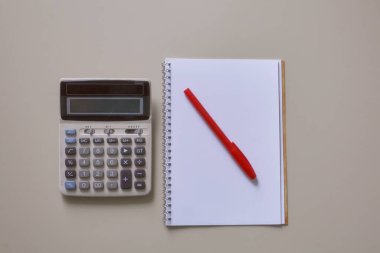 Masanın üzerinde bir defter, kalem ve hesap makinesi var. Kapı kolu kırmızı. Pastel gözlük. Yatay çerçeve.