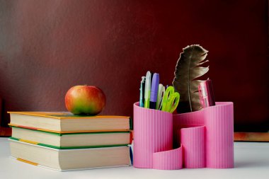 Masanın üzerinde kitaplar var. Kitapların yanında kalemlerle dolu bir kalem kutusu var. Arka planda bir karatahta var. Tahta kahverengi. Yatay çerçeve.