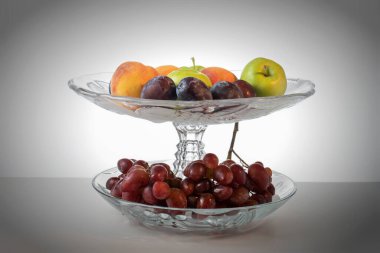 Masanın üzerinde bir meyve vazosu var. Vazoda üzüm ve şeftali var. Elmalar, erikler. Durgun hayat. Yatay çerçeve.