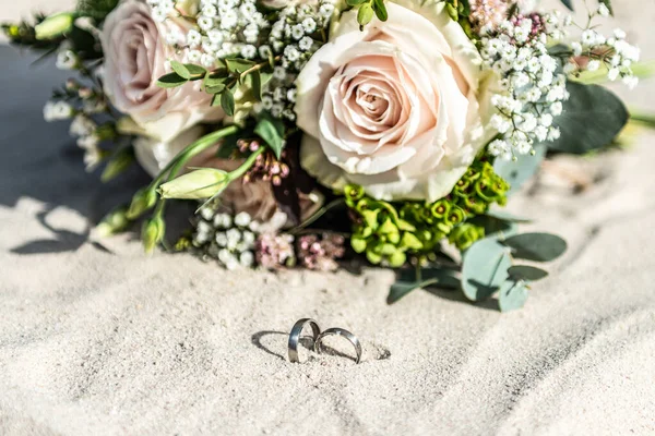 Os anéis de casamento bonitos encontram-se na superfície da areia na praia contra o fundo de um buquê de flores da noiva — Fotografia de Stock