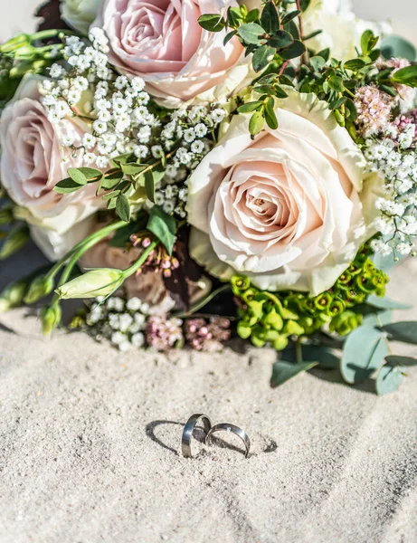 Красивые обручальные кольца лежат на песчаной поверхности на пляже на фоне букета цветов невесты — стоковое фото