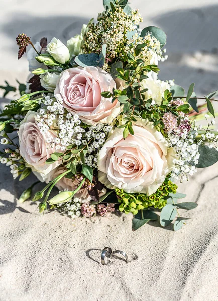 Hermosos anillos de boda se encuentran en la superficie de arena en la playa sobre el fondo de un ramo de flores novia — Foto de Stock