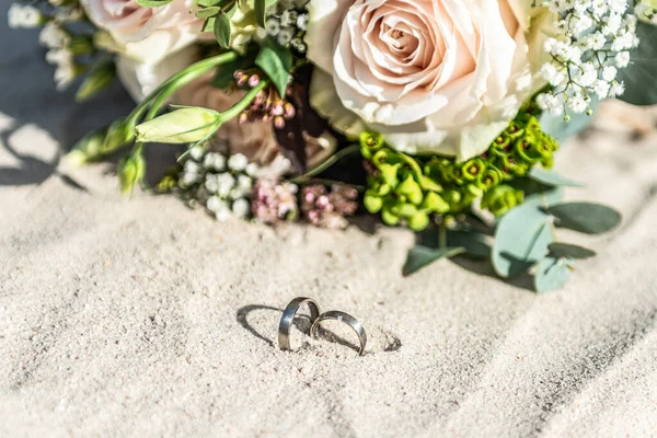 Hermosos anillos de boda se encuentran en la superficie de arena en la playa sobre el fondo de un ramo de flores novia — Foto de Stock