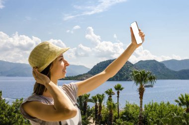 Gülümseyen kadın rahat sarı şapka smartphone deniz beach palm dağ güneşli bir gün bir arka plan üzerinde tutarak yaz tatili portresi