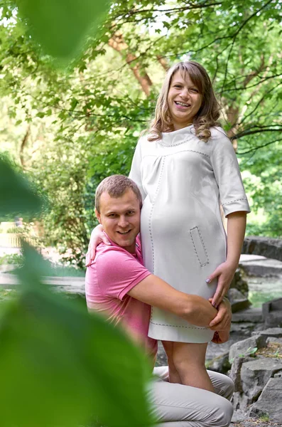 ハンサムな男は 彼の美しい妊娠中の妻のおなか腹を聞いて 笑顔します 夫は彼の妻の腹を聴きます 幸せな妊娠家族面白い — ストック写真