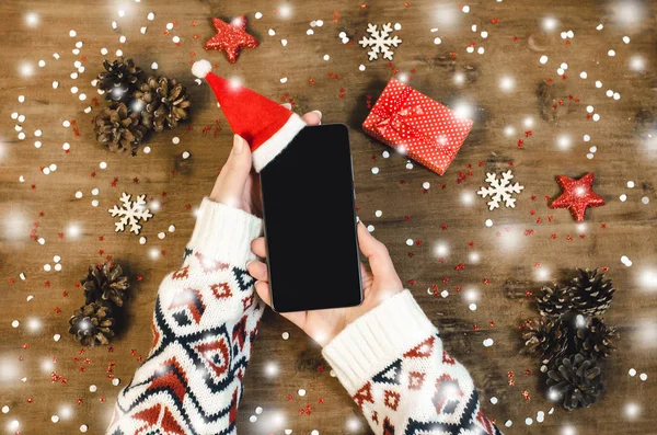 Female hands holding smart mobile phone with oled display on wooden background with Christmas gifts snowflakes and snow magic fairy tale light effect. Happy New Year and Xmas Flat lay composition top view.