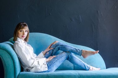 Gülümseyen genç kadın moda mavi kanepe stüdyo photoshoot kot mavi yırtık kot beyaz gömlek giyen oldukça serin model bakmak
