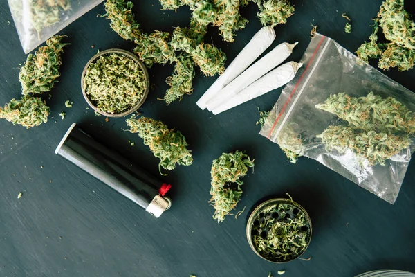 Bud of marijuana flowers on scales, grinder and shredded cannabis joint and a packet of weed and Smartphone on a black wood background — Stock Photo, Image