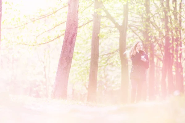 Parlant au téléphone, une fille marche le matin dans un parc avec un smartphone et un café, tonifiant la lumière — Photo