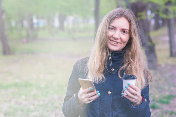 Una chica está caminando por la mañana en un parque con un teléfono inteligente y una luz de café tonificando — Foto de Stock