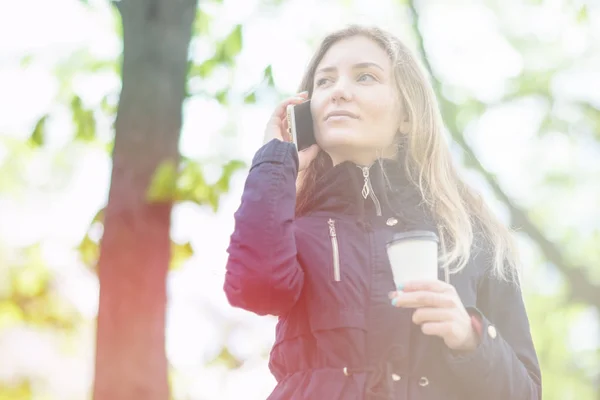 Hablando por teléfono, Una chica está caminando por la mañana en un parque con un teléfono inteligente y café, tonificación ligera — Foto de Stock