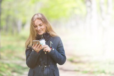 Sabah bir akıllı telefon ile bir parkta yürüyüş ve kahve, hafif tonlama bir kızdır