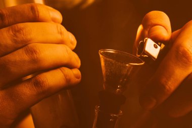 Bir adam bong esrar ot çekiyor. Siyah bir arka plan üzerinde duman. Sarı tonlama