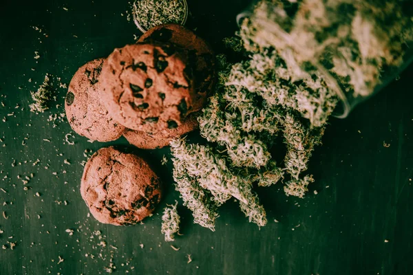 Galletas El chocolate con CBD y THC están sobre una mesa negra con cogollos de cannabis. El concepto de marihuana en la comida. Tratamiento con la ayuda de Canabis en un tono verde malhumorado . —  Fotos de Stock