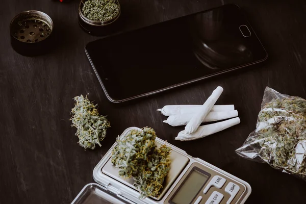 Los brotes de cannabis en la balanza se encuentran sobre la mesa con una amoladora, una articulación y un teléfono inteligente. Marihuana en el paquete. Fondo oscuro. De cerca. Color de tonificación —  Fotos de Stock