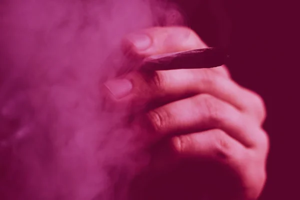 Een man rookt wiet, een joint en een aansteker in zijn handen. Rook op een zwarte achtergrond. Concepten van medicinaal marihuanagebruik en legalisering van de cannabis. Op een zwarte achtergrond Violet tinten — Stockfoto