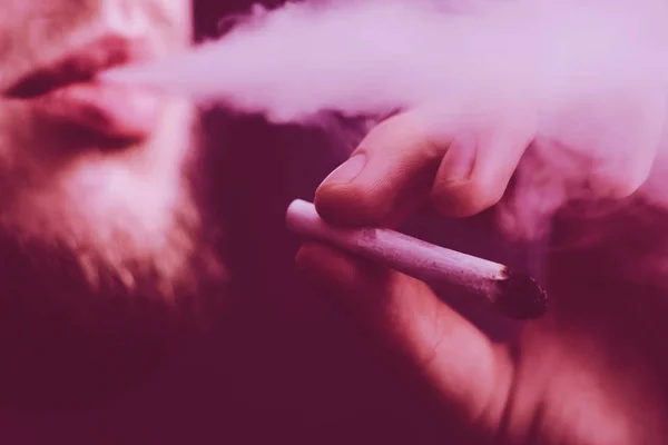 A joint in hand A man smokes cannabis weed, . Smoke on a black background. Concepts of medical marijuana use and legalization of the cannabis. On a black background Violet tinting — Stock Photo, Image