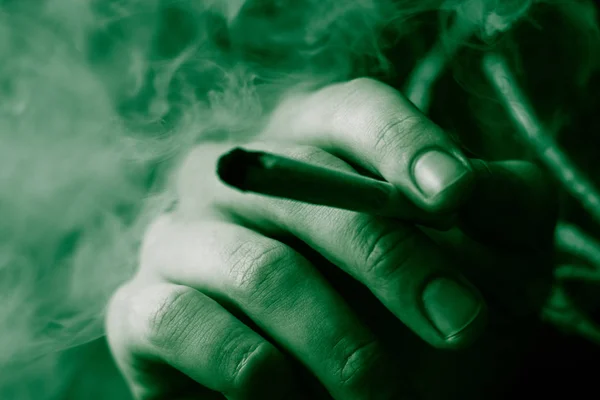 Erva de cannabis, um charro nas mãos Um homem fuma. Fumar em um fundo preto. Conceitos de uso medicinal de maconha e legalização da cannabis. Em um fundo preto Verde Tinting — Fotografia de Stock