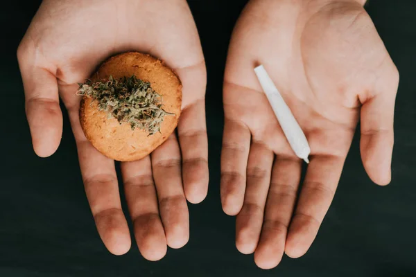 In den Händen eines Mannes Cannabis-Knospe und Joint und Kekse. das Konzept des Marihuanakonsums. auf schwarzem Hintergrund. Ansicht von oben . — Stockfoto