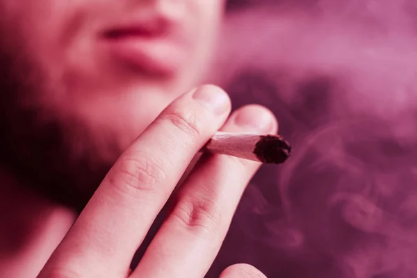 Humo sobre fondo negro. Un hombre fuma marihuana, un porro y un encendedor en sus manos. Conceptos de uso médico de la marihuana y legalización del cannabis. Sobre un fondo negro Tintado violeta — Foto de Stock