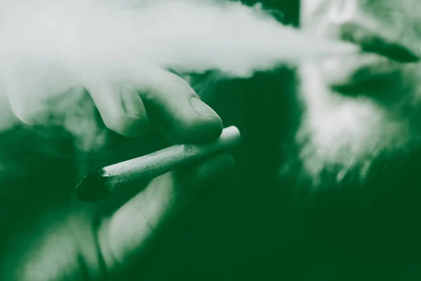 Un porro en la mano Un hombre fuma marihuana,. Humo sobre fondo negro. Conceptos de uso médico de la marihuana y legalización del cannabis. Sobre un fondo negro Tintado verde — Foto de Stock