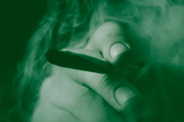 A man smokes cannabis weed, a joint and a lighter in his hands. Smoke on a black background. Concepts of medical marijuana use and legalization of the cannabis. On a black background Green Tinting — Stock Photo, Image