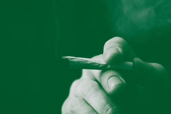 Un hombre fuma marihuana, un porro y un encendedor en sus manos. Humo sobre fondo negro. Conceptos de uso médico de la marihuana y legalización del cannabis. Sobre un fondo negro Tintado verde — Foto de Stock