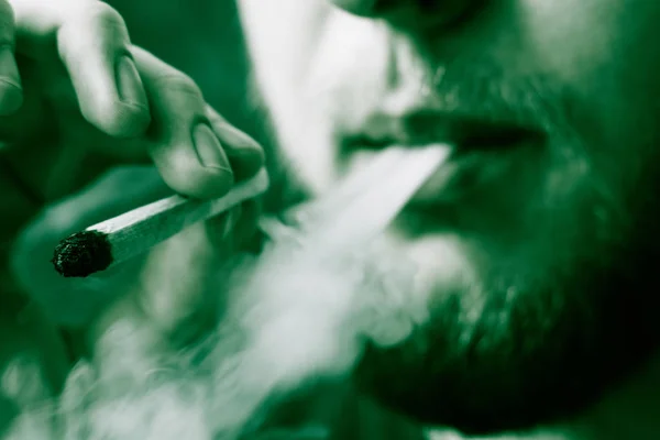 Ένας άνθρωπος καπνίζει κάνναβη ζιζανίων, ένα κοινό και ένα ελαφρύτερο στα χέρια του. Καπνού σε μαύρο φόντο. Έννοιες της ιατρική χρήση μαριχουάνα και τη νομιμοποίηση της κάνναβης. Σε μαύρο φόντο πράσινο χρωματισμό — Φωτογραφία Αρχείου
