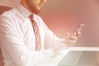 Finansal işler. Bilgisayarla ofis işi. Ofisteki masada bilgisayarla çalışan bir iş adamı. Hafif arkaplan. Kapatın. Elinde akıllı telefon Genç sakallı iş adamı.