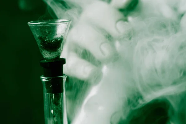 En man röker cannabis ogräs, en bong och en tändare i handen. Rök på en svart bakgrund. Begreppet medicinsk marijuana användning och legalisering av cannabis. På en svart bakgrund grön färgning — Stockfoto