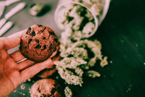 Cookies de chocolate com CBD e THC na mão estão em uma mesa preta com botões de cannabis. O conceito de maconha na comida. Tratamento com a ajuda de Canabis em um tom verde mal-humorado — Fotografia de Stock