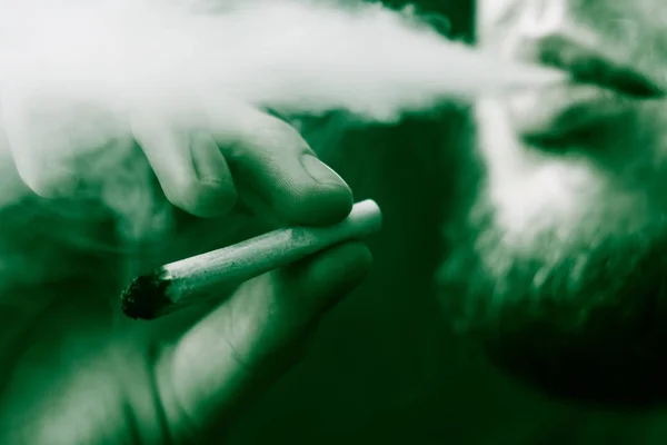 Koncepce užívání lékařské marihuany a legalizace konopí. Kouř na černém pozadí. Muž kouří konopí plevel, společné a zapalovač ve svých rukou. Na černém pozadí zelené tónovací — Stock fotografie
