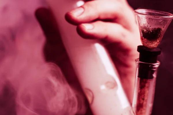 Fogalmak és a kannabisz legalizálása orvosi marihuána. Egy férfi dohányzik gyom kannabisz, egy bong, és egy könnyebb a kezében. A füst a fekete háttér. A fekete háttér, lila tónusokkal — Stock Fotó