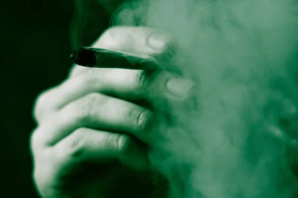 大麻杂草, 在他手中的一个男人抽烟的联合。黑色背景下的烟雾。医疗用大麻的概念和大麻的合法化。在黑色背景绿色着色 — 图库照片