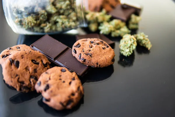 Chocolate e biscoitos com maconha e botões de maconha na mesa. Tratamento de maconha medicinal para uso em alimentos, fundo preto. Conceito de Cookies e chocolate com erva de cannabis CBD . — Fotografia de Stock