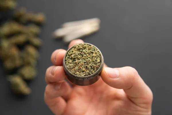 Cannabis buds on black table, joint with weed, top view close up, grinder with fresh marijuana in hand, — Stock Photo, Image