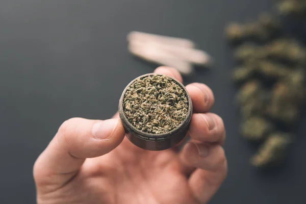Grinder with fresh marijuana in hand, top view close up, Cannabis buds on black table, joint with weed, — Stock Photo, Image