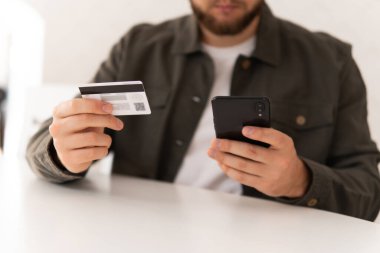 Kredi kartıyla internet alımı ve geçmişi bulanık bir şekilde evde oturan akıllı bir telefon. İnternetten alışveriş için el kredi kartı. İnternet alışverişi. İş yerinde online alışveriş akıllı telefonu ödemesi.
