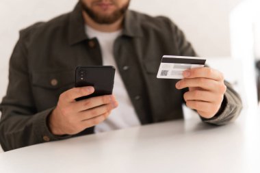 Kredi kartıyla internet alımı ve geçmişi bulanık bir şekilde evde oturan akıllı bir telefon. İnternetten alışveriş için el kredi kartı. İnternet alışverişi. İş yerinde online alışveriş akıllı telefonu ödemesi.