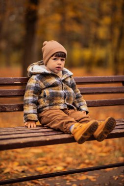 Kalın elbiseli küçük çocuk sonbahar parkında bankta oturuyor. Sonbahar konsepti, sonbahar havası.